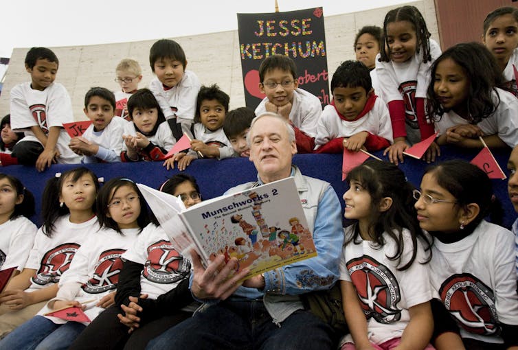 A man holds a storybook and he’s surrounded by children.